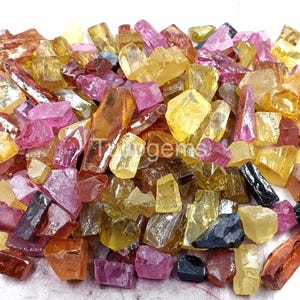 Puede incluir: Un montón de piedras preciosas en bruto de varios colores, incluyendo amarillo, rosa, naranja y negro. Las piedras preciosas tienen formas irregulares y parecen sin cortar. La imagen está bien iluminada, resaltando los diferentes colores y texturas de las piedras.