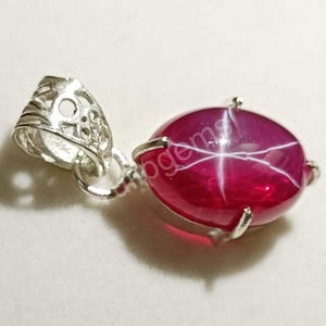 Può includere: Un pendente di rubino con effetto stella. La gemma ovale, rosso intenso, è incastonata in una montatura in metallo argentato. Il pendente ha un'ansa decorativa in argento.