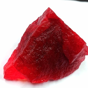 May include: A raw, uncut, ruby-red gemstone with a rough, textured surface. The stone is irregularly shaped, with sharp edges and a crystalline appearance. The gemstone is set against a white background.