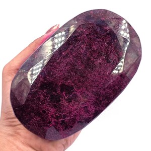 May include: A large, oval-shaped gemstone with a deep burgundy interior and a faceted, clear exterior. The gemstone is held in a hand, showcasing its size and color. The stone's surface reflects light, highlighting its intricate internal structure.