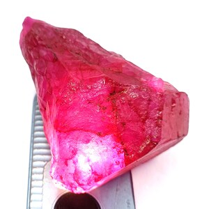 Puede incluir: Una gema de rubí en bruto, sin cortar, que muestra un tono rosa intenso y vibrante. La superficie rugosa de la piedra es visible, con texturas y variaciones naturales. El rubí está colocado sobre una herramienta de medición plateada, sobre un fondo blanco.