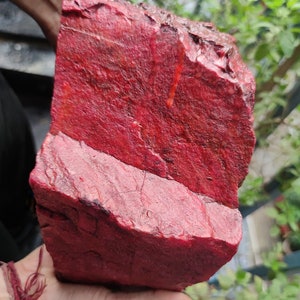 Puede incluir: Una gran roca en bruto con un tono rojo intenso. La piedra tiene una apariencia en capas, con una banda de color más claro que atraviesa horizontalmente el centro. La superficie es texturizada e irregular, con grietas y variaciones de color visibles.