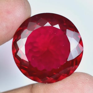 May include: A round, faceted gemstone with a deep, vibrant red color. The stone features a brilliant cut with numerous triangular and kite-shaped facets that reflect light, creating a clear, sparkling appearance across its circular surface.