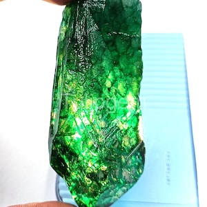 Op de afbeelding: Een ruwe, diepgroene smaragd edelsteen. De onbewerkte smaragd vertoont een kristallijne structuur met verschillende groentinten en een reflecterend oppervlak. De edelsteen wordt tussen de vingers gehouden, wat de natuurlijke vorm laat zien. De smaragd is een natuurlijke edelsteen.