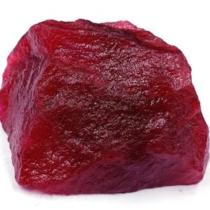 May include: A rough-cut, raw ruby gemstone. The ruby is a deep, saturated red color with a textured surface. The gemstone is irregularly shaped and appears to be unpolished, showcasing its natural form. This item is suitable for jewelry making or gemstone collecting.