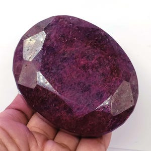 Puede incluir: Una gran gema de rubí ovalada y facetada. La gema es de un color burdeos intenso con facetas reflectantes. La piedra se sostiene en una mano sobre un fondo blanco.