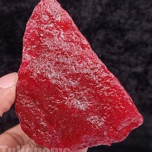 May include: A rough-cut, vibrant red gemstone with a textured surface. The gemstone is held by a hand, with the text "Tulipgems" visible at the bottom. The stone's color is a deep, saturated red.