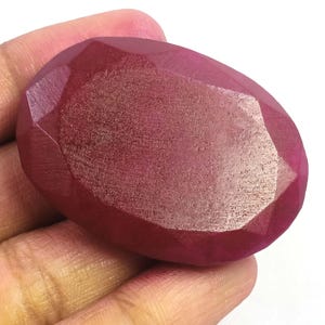Peut inclure: Une pierre précieuse rubis facettée de forme ovale. La gemme est d'un rouge profond et riche avec de multiples facettes qui captent la lumière. La pierre est tenue dans une main, mettant en valeur sa taille et sa forme. Cette pierre précieuse est adaptée à la joaillerie.