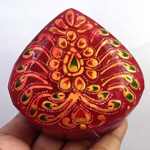 Puede incluir: Una caja roja en forma de corazón con un diseño pintado a mano que presenta motivos en forma de lágrima amarillos y verdes y pequeños puntos blancos. La superficie tiene un acabado brillante y la tapa está decorada con patrones simétricos.