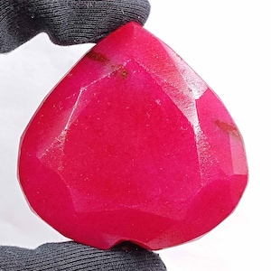 May include: A heart-shaped, faceted gemstone in a vibrant red hue. The gem features multiple flat surfaces that reflect light, creating a sparkling effect. The stone is held by a black glove.