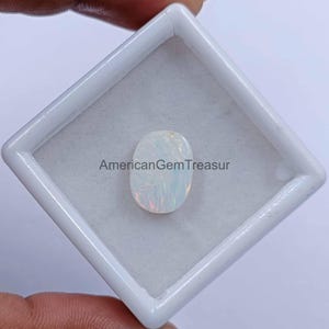 May include: An oval-shaped opal gemstone, set in a white plastic case. The opal has a milky white base color with flashes of iridescent colors. The text "AmericanGemTreasur" is printed on the case.