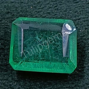 Puede incluir: Una gema rectangular con corte esmeralda de un tono verde vibrante. La gema tiene una apariencia transparente con inclusiones internas visibles y una superficie pulida. Los bordes están cortados con precisión, mostrando la claridad y forma de la gema.