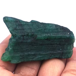 May include: A raw, uncut emerald gemstone with a deep green color. The rough stone has a textured surface with visible lines and ridges. The emerald is held in a hand, showcasing its natural form and color.