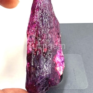 May include: A raw, uncut gemstone with a deep purple and magenta color. The rough surface of the stone has a textured appearance. The gemstone is held up against a light gray surface.