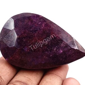 Puede incluir: Una gema facetada en forma de pera con un tono púrpura intenso. La gema tiene una superficie pulida y una faceta tallada en la parte superior izquierda. El texto "Tulipgem" es visible. La gema se sostiene en una mano.