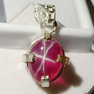 Pode incluir: Pingente de prata com uma grande pedra preciosa ovalada, vermelho escuro. A pedra preciosa tem um padrão em forma de estrela em sua superfície. O pingente tem uma argola decorativa e é fixado em uma moldura de metal prateado com detalhes dourados.