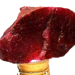 May include: A large, rough-cut ruby gemstone with a deep red color. The surface has a textured appearance, and the stone is illuminated from below, highlighting its rich hue. This natural gemstone is a striking example of raw mineral beauty.