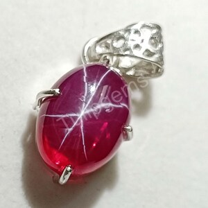 May include: A ruby pendant with a star effect. The oval-shaped, deep red gemstone is set in a silver-colored metal setting. The pendant has a textured bail for a chain.