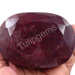 Puede incluir: Una gran gema de rubí de forma ovalada y multifacética. La gema es de color rojo intenso con una superficie pulida. El texto "Tulipgems" está impreso en la superficie.