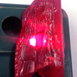 May include: A raw, translucent ruby gemstone with a deep red hue. The rough-cut stone has a textured surface and a bright, internal glow. The ruby is displayed against a dark green background.