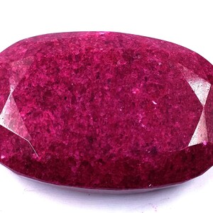 Puede incluir: Una gema de rubí facetada, de forma ovalada. La gema es de un color rojo intenso y saturado con una superficie pulida. Las facetas capturan la luz, creando reflejos y sombras. La gema se encuentra sobre un fondo blanco.