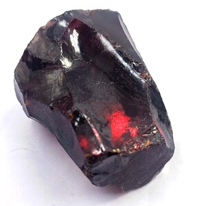 May include: A raw, uncut gemstone with a deep burgundy color and a glossy, reflective surface. The stone has a rough, irregular shape, with facets catching the light. The overall appearance is dark and rich, with hints of red.