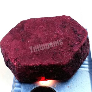 Puede incluir: Una gran gema de rubí en bruto, de color rojo intenso. La piedra tiene forma hexagonal y una superficie texturizada. El texto "Tulingems" es visible en la superficie de la piedra.
