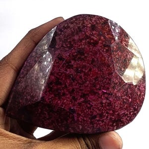 Puede incluir: Una gema grande y facetada de color burdeos intenso con un patrón moteado. La piedra se sostiene en una mano, mostrando su tamaño y textura única. El corte crea múltiples superficies reflectantes.