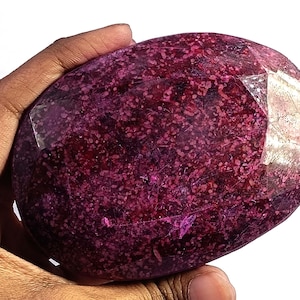 May include: A large, faceted gemstone with a deep magenta hue, held in a hand. The stone's surface is speckled with lighter pink inclusions, and it has a polished, oval shape with multiple facets that catch the light.