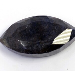 Puede incluir: Una gema facetada de color azul oscuro con corte marquesa. La gema tiene una superficie pulida con múltiples facetas que reflejan la luz. La forma es alargada y puntiaguda en ambos extremos. La gema se encuentra sobre un fondo blanco.