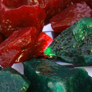Op de afbeelding: Close-up van ruw geslepen edelstenen in tinten rood en groen. De rode stenen hebben een glanzende, doorschijnende uitstraling, terwijl de groene stenen ondoorzichtig zijn. De edelstenen zijn onregelmatig van vorm en lijken ruwe, natuurlijke materialen te zijn.