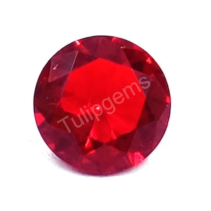 Può includere: Una gemma sfaccettata, rotonda, di colore rosso brillante. La gemma ha un taglio brillante con molteplici sfaccettature che riflettono la luce. Il testo "Tulipgems" è stampato sulla gemma.