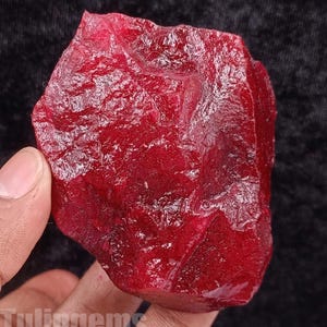 May include: A rough, uncut ruby gemstone with a deep red color. The surface of the stone has a textured, uneven appearance. The word "Tuligem" is visible at the bottom of the image.