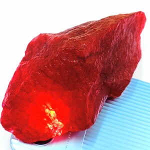 May include: A large, rough-cut ruby gemstone with a deep red color. The surface has a textured appearance, and the stone is illuminated from within, creating a bright glow. The gemstone is displayed on a light blue surface.