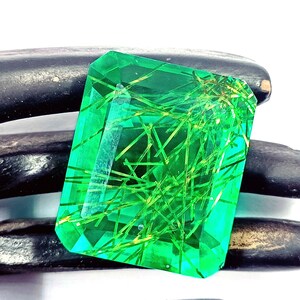 以下が含まれることがあります： 鮮やかなエメラルドカットの宝石で、鮮やかな緑色と内部の針状のインクルージョンが特徴です。 宝石は暗く質感のある背景に配置され、その透明度と輝きを際立たせています。 自然の鉱物芸術の美しい例です。