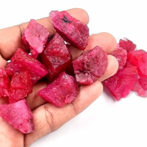 May include: A handful of rough-cut, vibrant red ruby gemstones. The raw, natural stones display a range of shapes and sizes, with a deep, saturated color. The gems are held in a hand against a white background.