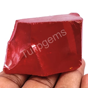 Może przedstawiać: Surowy, głęboko czerwony kamień szlachetny o lekko przezroczystym wyglądzie. Powierzchnia ma naturalną, nierówną fakturę. Tekst "Turfgems" jest widoczny. Kamień szlachetny jest trzymany w dłoni.