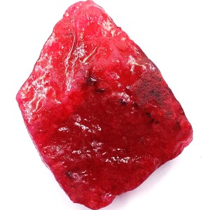 Pode incluir: Uma gema de rubi bruta, não cortada, com um tom vermelho vibrante. A superfície tem uma aparência ligeiramente texturizada com manchas mais escuras, contra um fundo branco limpo. Esta gema natural é um exemplo marcante de beleza mineral bruta.