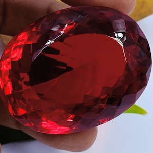 May include: A large, faceted, oval-shaped gemstone with a deep red hue. The gem is held between fingers, showcasing its polished surfaces and internal reflections. The cut of the stone creates numerous facets, enhancing its brilliance and color.