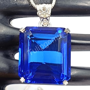 May include: A rectangular, deep blue gemstone pendant with a silver-colored bail and chain. The gemstone is faceted, catching the light and displaying a brilliant shine. The pendant is a striking piece of jewelry.