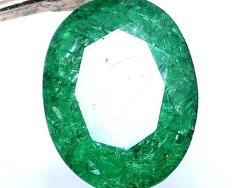 Pode incluir: Esmeralda de corte oval, segurada por pinças prateadas. A gema tem uma superfície facetada com uma cor verde brilhante e uma grande faceta geométrica branca no centro, sobre um fundo branco.