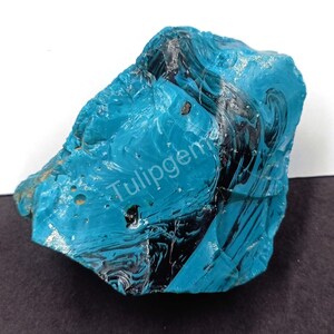 May include: A large, irregularly shaped, teal-colored stone with black veining. The stone has a rough, textured surface and appears to be a natural mineral specimen. The word "Tulipgems" is printed on the stone.