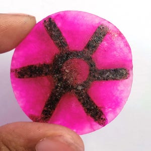 May include: A round, hot pink gemstone with a dark, geometric pattern. The pattern is a six-pointed star design with a central circle. The gemstone is held by a person's fingers.