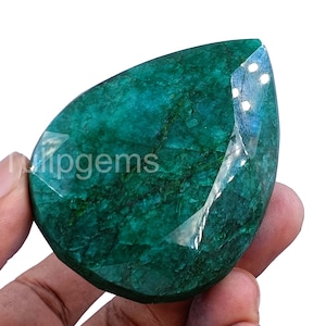Puede incluir: Una gran esmeralda facetada de color verde intenso. La gema tiene forma de lágrima y se sostiene en una mano, mostrando sus superficies pulidas y su claridad interna. El texto "Tulipgems" es visible en la gema.