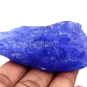 Puede incluir: Una gema de tanzanita en bruto, sin cortar, de color azul intenso. El mineral en bruto se sostiene en una mano, mostrando su textura natural y estructura cristalina. La palabra "tulipgem" es visible en la superficie.