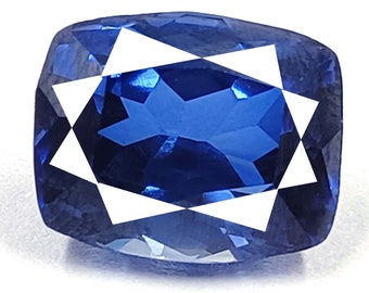 May include: A cushion-cut, deep blue gemstone with a faceted surface. The gem has a white geometric design overlaying the center. The stone is set against a white background, highlighting its color and cut.