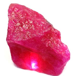 May include: A rough, uncut ruby gemstone. The ruby is a deep, vibrant red color with a textured surface. The gemstone is irregularly shaped and appears to be a natural, unpolished specimen. The ruby is set against a white background.