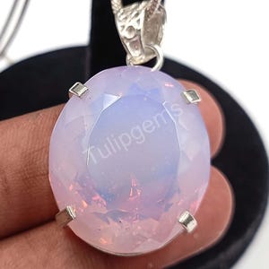 May include: A large, oval-cut pendant with a pale pink and lavender gemstone. The gemstone is set in a silver-colored metal with four prongs. The pendant hangs from a delicate silver chain. The text "tulipgems" is visible on the gemstone.