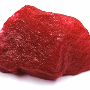 May include: A rough-cut, deep red gemstone with a textured surface. The stone has an irregular shape and is set against a white background. The color is a vibrant, saturated red.