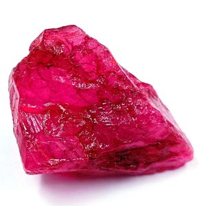 Puede incluir: Una gema de rubí roja en bruto. El rubí presenta una estructura cristalina y una superficie brillante. La gema se encuentra sobre un fondo blanco, resaltando su rico color y forma natural. Este rubí en bruto es un buen ejemplo de formaciones minerales naturales.